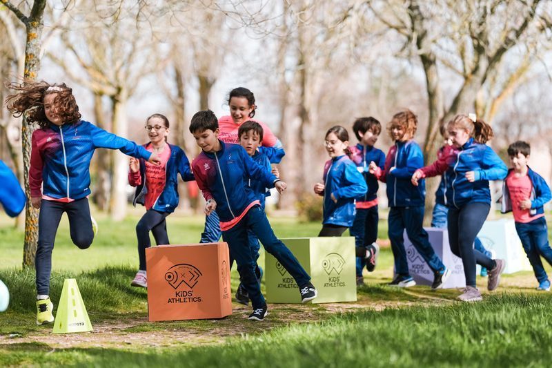 Niños jugando al aire libre, corriendo y saltando cajas, vistiendo chaquetas azules y rojas en un parque con césped.