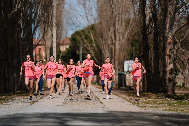 Un grupo de mujeres con camisetas rosas y pantalones cortos corriendo por un sendero bordeado de árboles.